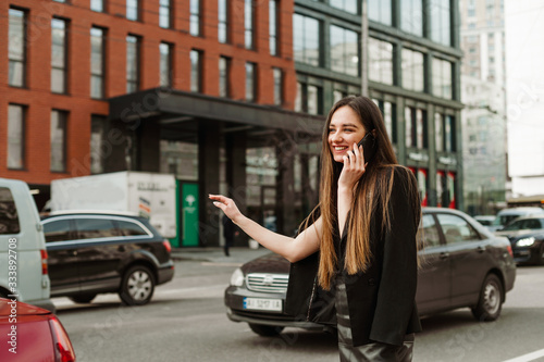 Happy busy businesswoman in casual clothes talking on the phone and catches a hand gesture. Smiling formal-dressed lady communicates on the phone and stops a taxi against a cityscape. © bodnarphoto