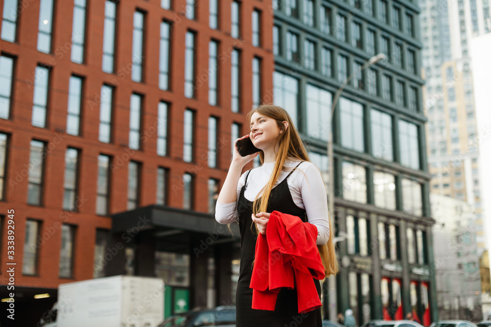 Smiling young girl in formal wear and red jacket in hands communicates on the phone against urban background, looks away and smiles. Lady student talking on the phone on the street. Smiling young girl in formal wear and red jacket in hands communicates on the phone against urban background, looks away and smiles. Lady student talking on the phone on the street.