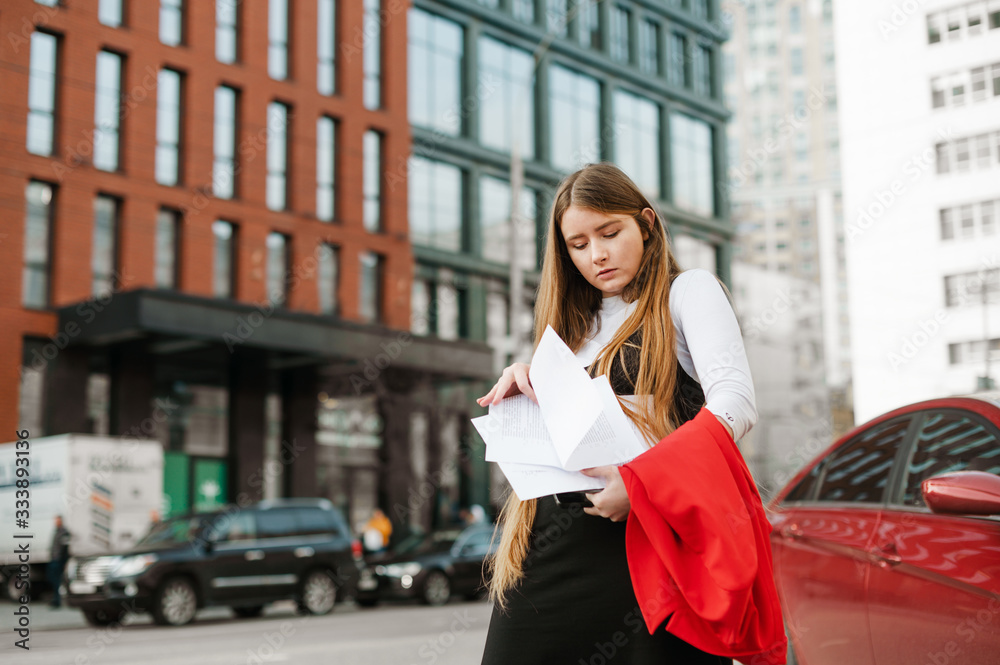 Portrait of busy student girl standing outdoors on cityscape background with laptop and notes in hands, confused looking for right piece of paper. Girl nerd is studying the way to the university.