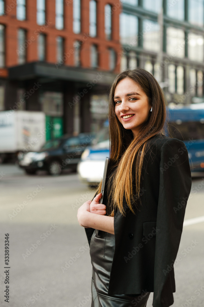 Pretty positive young businesswoman with long hair and in wireless headphones posing for the camera, holding a laptop, looking into the camera with a smile on her face. Happy business woman on street.