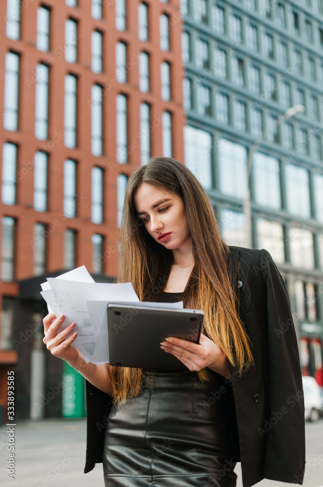 Portrait of attractive businesswoman in office clothes stands on cityscape background with laptop and papers in hands, focuses on business papers. Vertical photo. Business concept. Portrait of attractive businesswoman in office clothes stands on cityscape background with laptop and papers in hands, focuses on business papers. Vertical photo. Business concept.