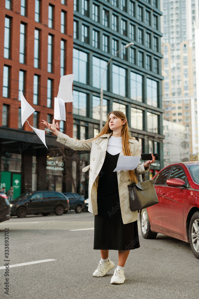 Girl with long hair stands on the street and throws up notes with a serious face, vertical photo. Young girl student throws away paper from anger on cityscape background.