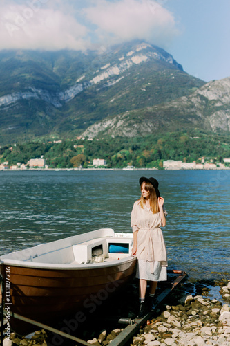  girl in a hat stands near a boat on the lake on a background of mountains