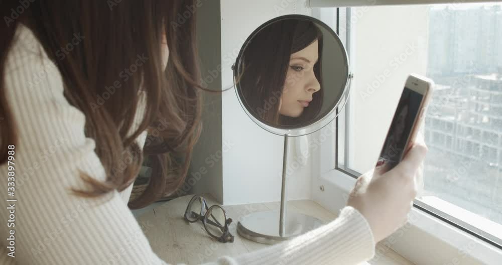 Taking selfie. Rear view of beautiful young woman taking self portrait in the mirror using smart phone while sitting on bed at home. Communication and socialization with new intellectual technologies. Taking selfie. Rear view of beautiful young woman taking self portrait in the mirror using smart phone while sitting on bed at home. Communication and socialization with new intellectual technologies.