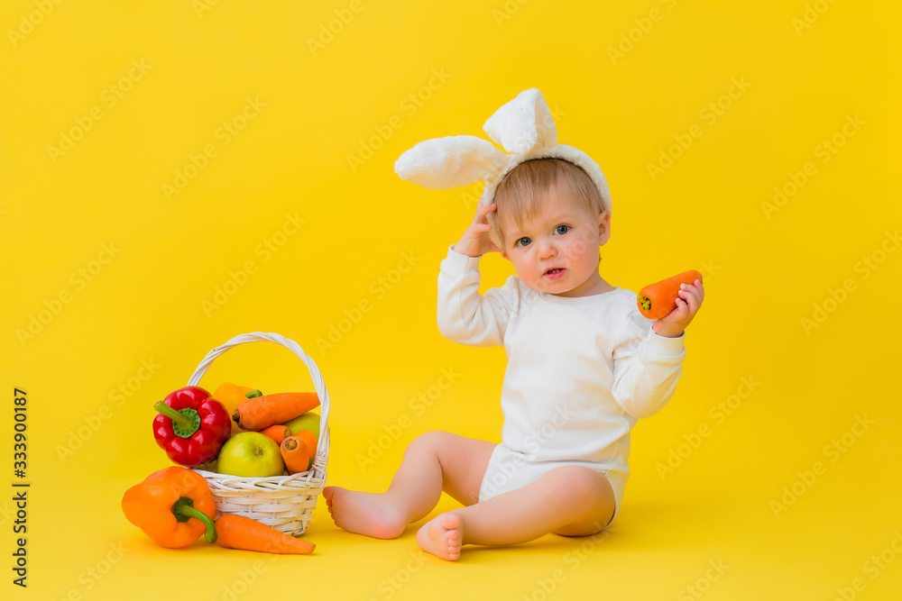 Baby in a white bodysuit with rabbit ears on his head eats carrots, sits on a yellow background with vegetables. baby in the form of an Easter bunny, space for text Baby in a white bodysuit with rabbit ears on his head eats carrots, sits on a yellow background with vegetables. baby in the form of an Easter bunny, space for text