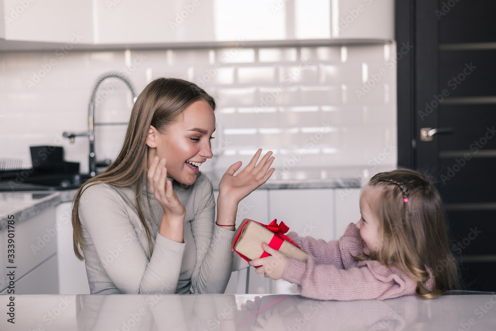Cute young daughter offering gift to her mother in the in the kitchen. Mother day. Cute young daughter offering gift to her mother in the in the kitchen. Mother day.