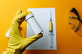 Doctor in yellow gloves holds coronavirus vaccine in white medicine jar, pills for covid-19 curing. Yellow syringe, protective glasses and list of infected people on orange background.