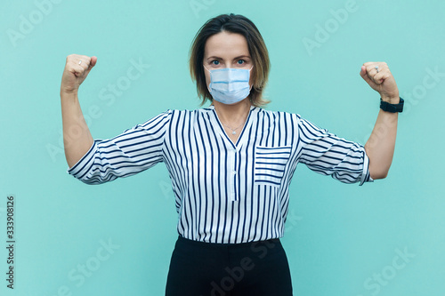 I am strong and I will be healthy. Portrait of proud satisfied middle aged woman with surgical medical mask standing with raised arms. medicine, health care concept. indoor isolated on blue background photo