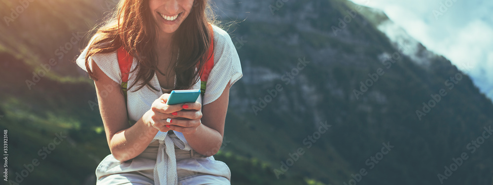Young woman sitting on the fence with mobile phone. High mountains touristic path at sunset. Texting with friends. Wide screen panoramic - obrazy, fototapety, plakaty 