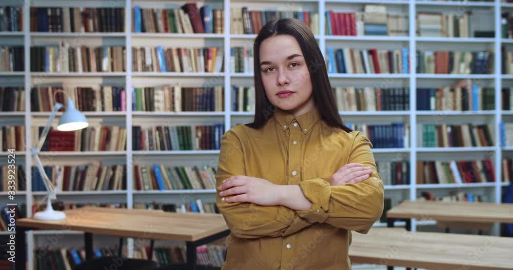 Portrait of talented young brunette girl crossing her arms and looking at the camera with angry face expression. Bookcase in background. Portrait of talented young brunette girl crossing her arms and looking at the camera with angry face expression. Bookcase in background.