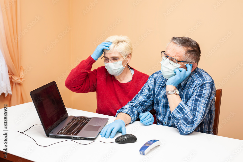 A pair of elderly people in medical masks and gloves are sitting at a table with a laptop