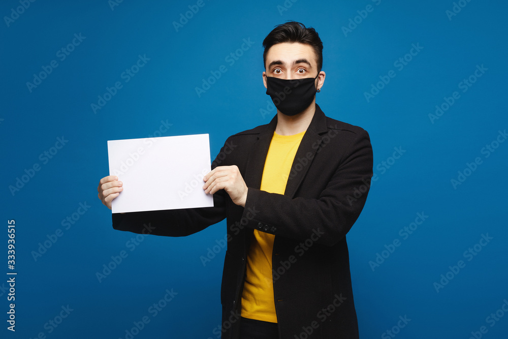 Young shocked man in a black protective mask holding an empty sheet of paper and looking in the camera, isolated at the blue background. Promotion concept. Healthcare concept Young shocked man in a black protective mask holding an empty sheet of paper and looking in the camera, isolated at the blue background. Promotion concept. Healthcare concept