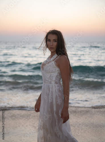 Romantic portrait of beautiful brunette woman in white lace dress standing at the beach over sea and sunset sky background