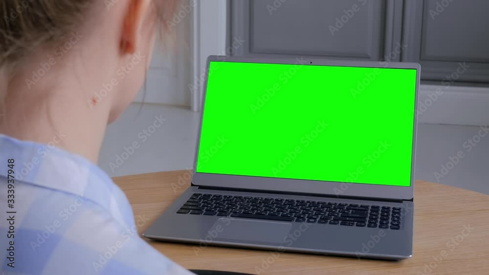 Woman looking at grey laptop with blank green display on wooden table in home interior. Mock up, copyspace, freelance workspace, chroma key, template, entertainment, green screen, technology concept Woman looking at grey laptop with blank green display on wooden table in home interior. Mock up, copyspace, freelance workspace, chroma key, template, entertainment, green screen, technology concept
