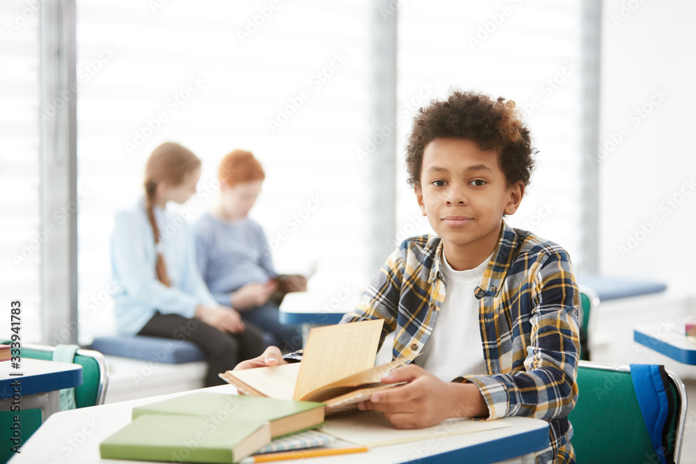 Portrait of cute African-American boy looking at camera while sitting at desk in school classroom, copy space - obrazy, fototapety, plakaty 
