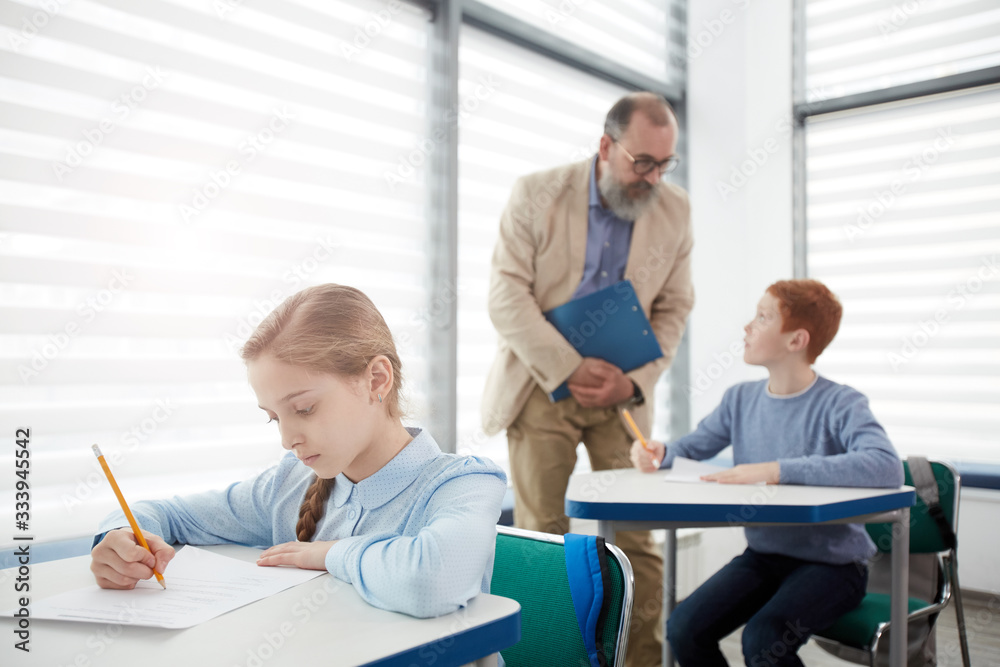 Group of children taking test with bearded mature teacher watching them, focus on cute blond girl sitting at desk and writing in foreground, copy space