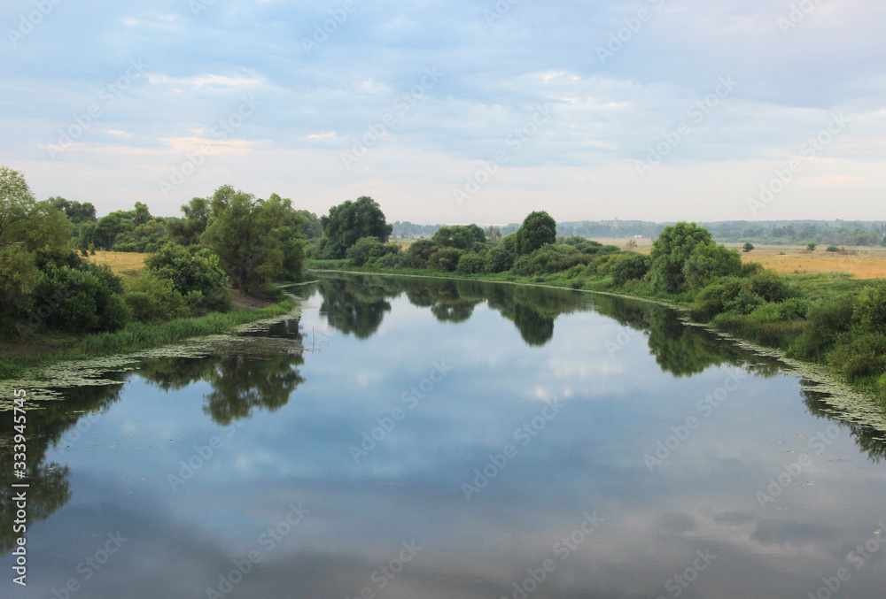 A wide river on a bend with reflected clouds in its surface and surrounded by trees and bushes.