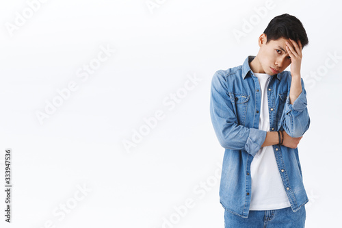 Portrait of tired and annoyed young asian man feeling burnout and huge stress, facepalm, look from under forhead with judgemental tired expression, cant stand person telling nonsense photo