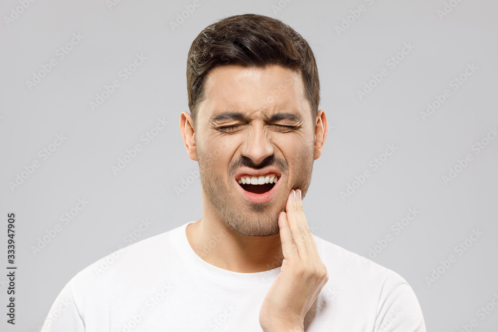Toothache concept. Young man touching his chin and closing eyes with expression of suffer from tooth ache, isolated on gray background