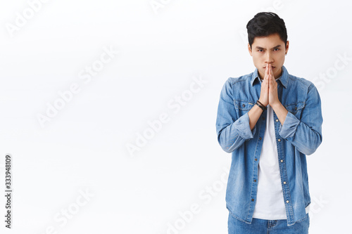 Portrait of worried, serious-looking handsome asian man praying, looking concerned as waiting important news, hold hands in pray, pleading, standing hopeful over white background