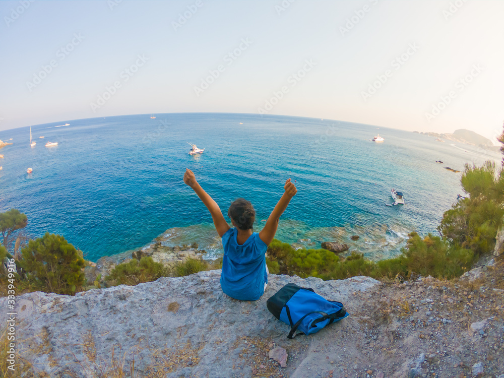 Young woman with backpack sitting on a cliff looking at the ocean spreading arms. Ponza Island coast, Italy. Young woman with backpack sitting on a cliff looking at the ocean spreading arms. Ponza Island coast, Italy.