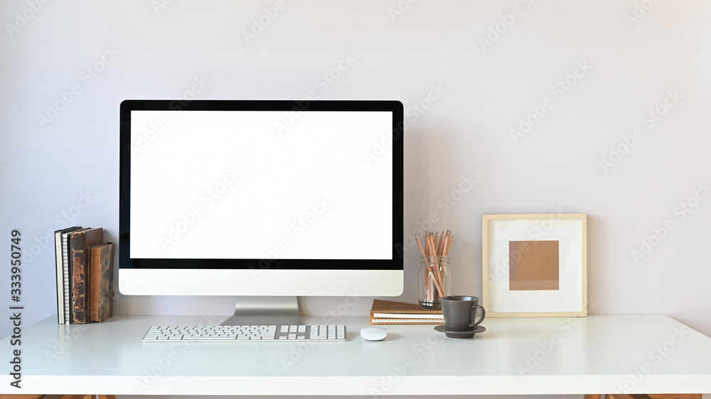 Workspace computer monitor with white blank screen putting on white working desk with wireless keyboard, mouse, coffee cup, picture frame, books and pencil holder over white wall as background. Workspace computer monitor with white blank screen putting on white working desk with wireless keyboard, mouse, coffee cup, picture frame, books and pencil holder over white wall as background.