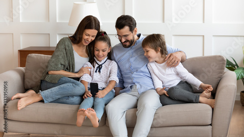 Front view joyful little child girl holding cellphone, showing funny video to laughing parents and brother, resting on sofa. Happy couple with children using funny mobile apps, enjoy weekend time. © fizkes