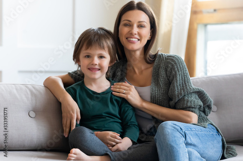 Portrait of smiling 30s woman cuddling small cute child boy, sitting together on sofa. Happy young mother hugging little adopted preschool kid son, looking at camera, posing for family photo.