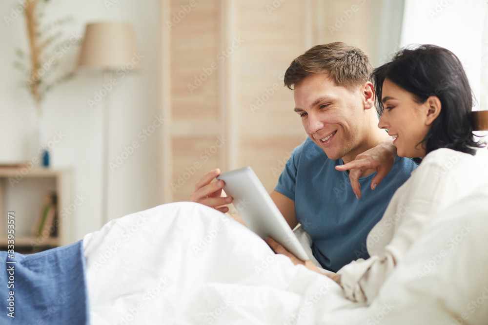 Side view portrait of smiling adult couple using digital tablet in bed in home interior while choosing online services, copy space Side view portrait of smiling adult couple using digital tablet in bed in home interior while choosing online services, copy space
