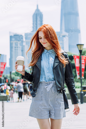 Happy young trendy red hair woman wearing black leather jacket and shorts, drinking take away coffee and walking in an urban city. City walk lifestyle concept.