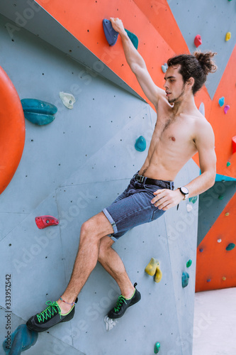 Urban concept of man with curly long hair, tied in a ponytail, is training at the city artificial red and blue climbing wall using talcum powder, wearing a bag for climbing without insurance equipment
