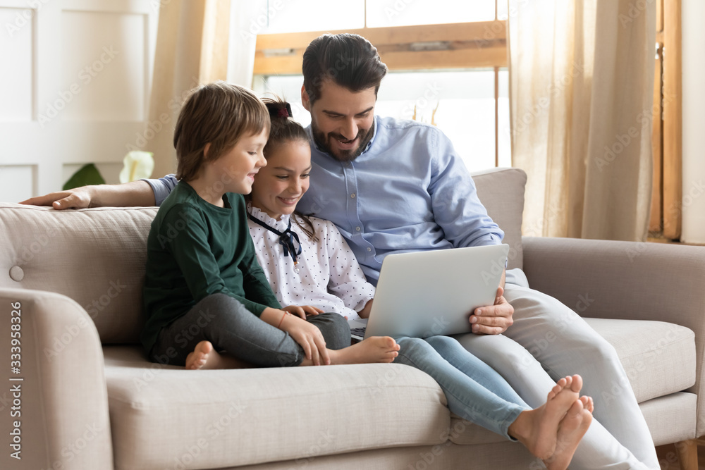 Smiling young dad resting on couch with cute children, showing educational applications on computer. Happy man enjoying spending free leisure time with kids son daughter, watching movie at home. Smiling young dad resting on couch with cute children, showing educational applications on computer. Happy man enjoying spending free leisure time with kids son daughter, watching movie at home.