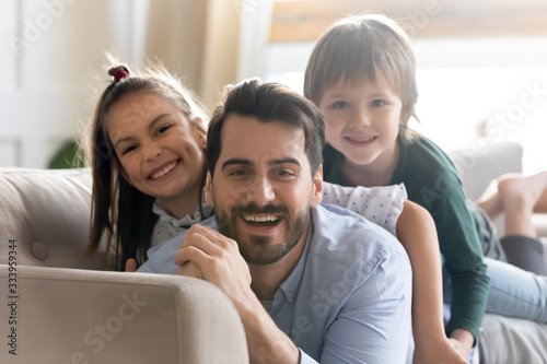 Head shot close up smiling young dad lying on sofa with adorable small children siblings, looking at camera. Happy bearded father holding on back playful little kids, posing for photo on couch.
