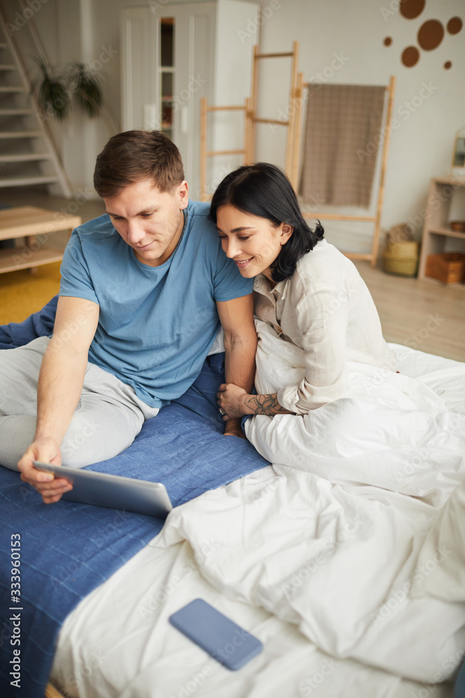 Vertical high angle portrait of young happy couple using digital tablet while sitting on bed at home and ordering food online or watching movie, copy space Vertical high angle portrait of young happy couple using digital tablet while sitting on bed at home and ordering food online or watching movie, copy space