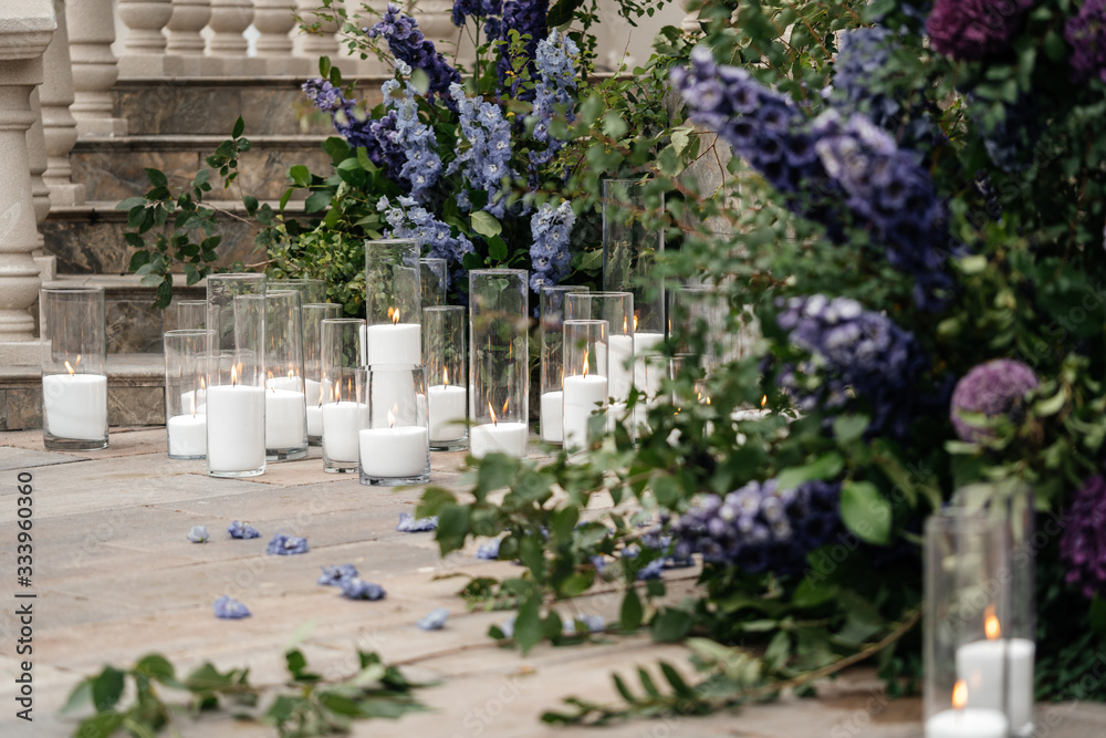 Burning white candles in glass vases on the floor near the greenery and flowers. Details of the wedding ceremony.
