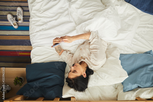 Top view portrait of modern young woman using smartphone while lying on bed in morning, copy space