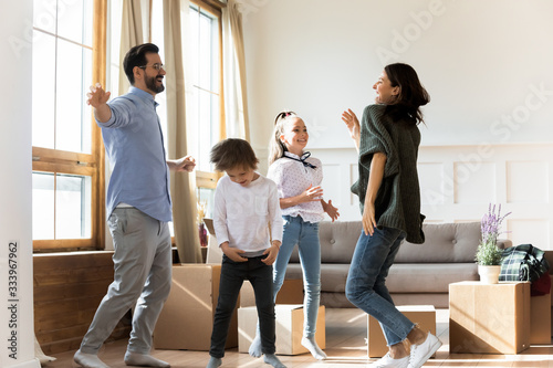 Full length overjoyed young parents dancing to energetic music with small kids siblings near cardboard boxes in new apartment. Excited homeowners celebrating moving in house flat, real estate concept.
