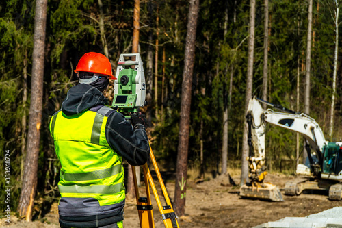 Surveyor engineer with equipment (theodolite or total positioning station) on the construction site of the road or building with construction machinery background