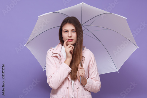 Horizontal photo of thoughtful attentive attractive female holding umbrella, putting one finger on cheeck, having pensive facial expression, wearing casual clothes. Weather forecast concept. photo