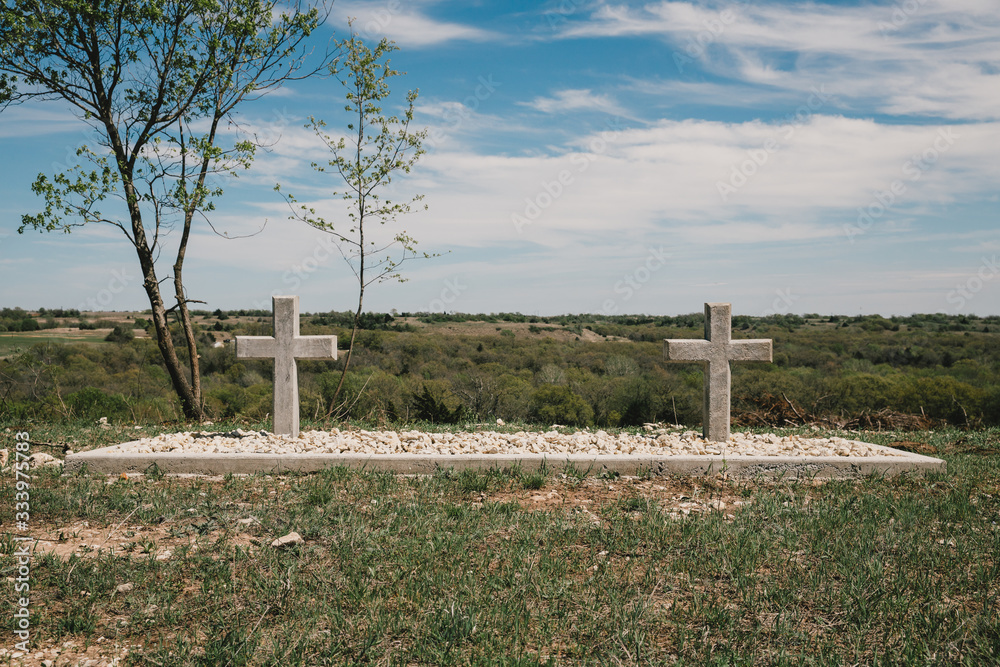 Two concrete cross shaped headstone grave markers stand side by side at the top of a hill overlooking the horizon to commemorate the lives of husband and wife