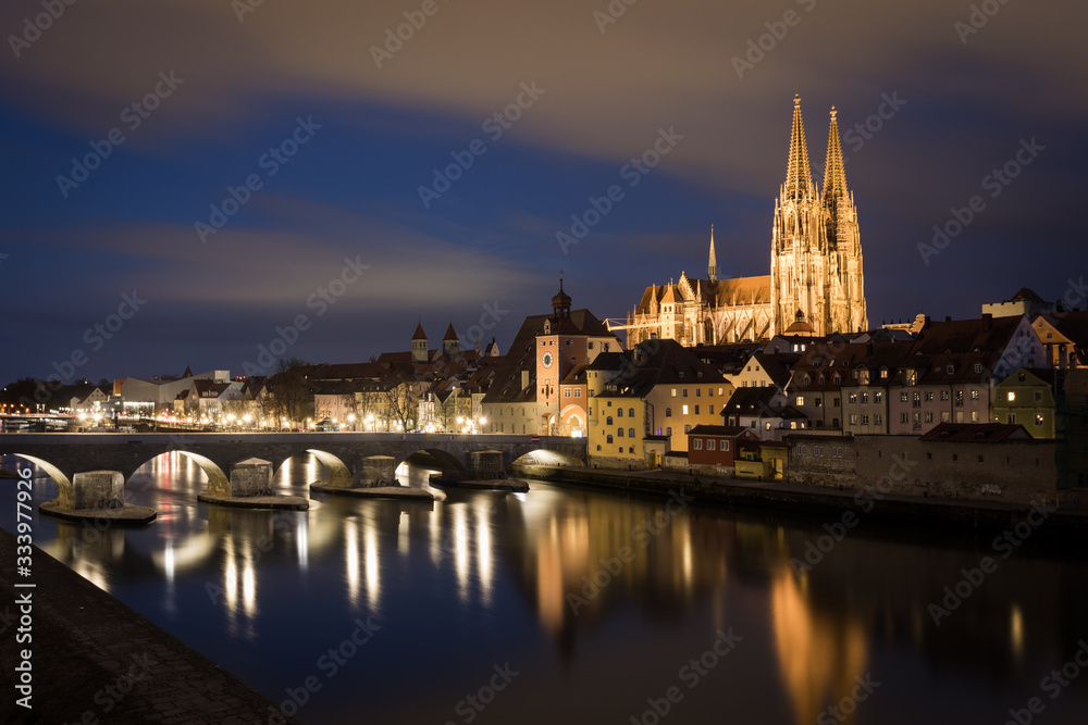 Illuminated saint Peter cathedral with historical stone bridge after dark in Regensburg, Bavaria, Germany. Cityscape image over the Danube river from Sorat Hotel terrace.