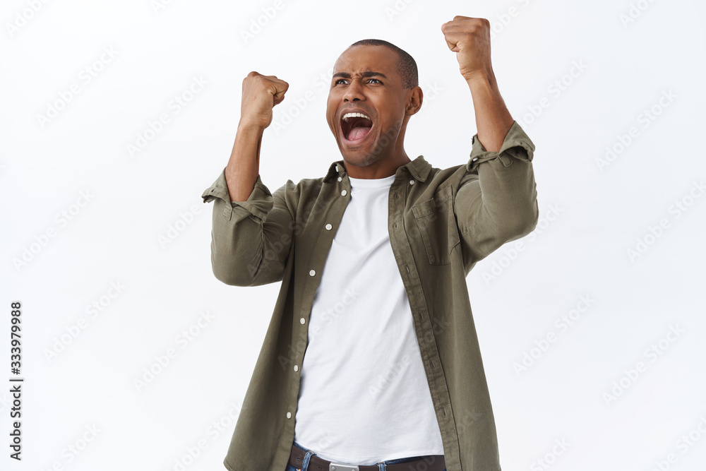 © Cookie Studio - Portrait of happy, triumphing african-american man celebrating victory, football fan winning bet in sports game, raising hands up cheerful, rooting for team, achieve success, white background