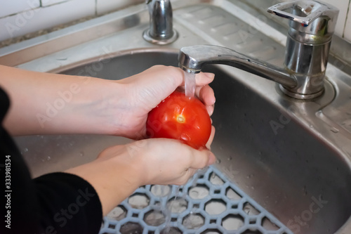Girl is washing bright red tomato under pouring water in the sink in the kitchen. Concept of hygiene, cleanliness and health protection