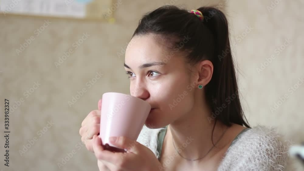 A young attractive girl is drinking coffee from a pink cup, smiling and looking at the camera. A young, sympathetic girl is enjoying aromatic coffee while sitting on the bed at home. A young attractive girl is drinking coffee from a pink cup, smiling and looking at the camera. A young, sympathetic girl is enjoying aromatic coffee while sitting on the bed at home.