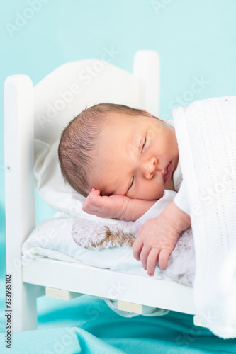 Little newborn baby boy falling asleep in tiny white bed on blue background. Cute new born sleeping during studio photoshoot. Safe sleep, sweet dreams concept.