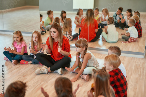 Having some fun after choreography class. Young female dance teacher with group of happy children clapping and playing while sitting on the floor in dance studio