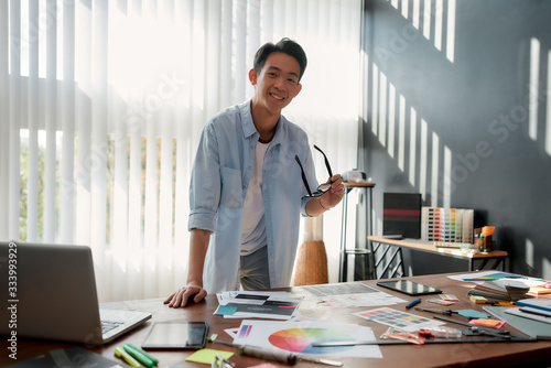 Happy at work. Cheerful asian man holding eyeglasses and smiling at camera while leaning on a table with a lot of creative stuff on it. Young designer in casual wear working in the modern office