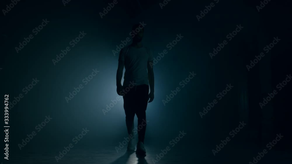 Young man in sports wear walking through smoky studio or scene directly to camera. Backlight, bright spotlight on background. Young man in sports wear walking through smoky studio or scene directly to camera. Backlight, bright spotlight on background.