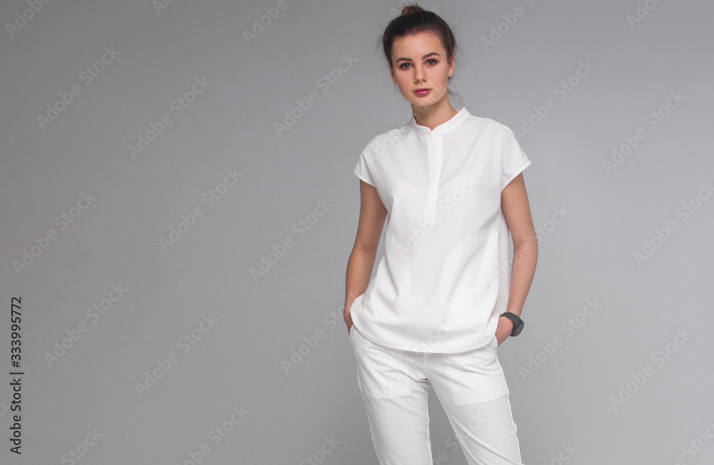 young stylish brunette nurse with long legs in white medical costume is standing with hands in her pockets, looking straight and smiling on gray background. medical concept. free space on left side young stylish brunette nurse with long legs in white medical costume is standing with hands in her pockets, looking straight and smiling on gray background. medical concept. free space on left side
