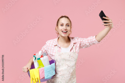 Excited young woman housewife in apron hold basin with detergent bottles washing cleansers while doing housework isolated on pink background. Housekeeping concept. Doing selfie shot on mobile phone.